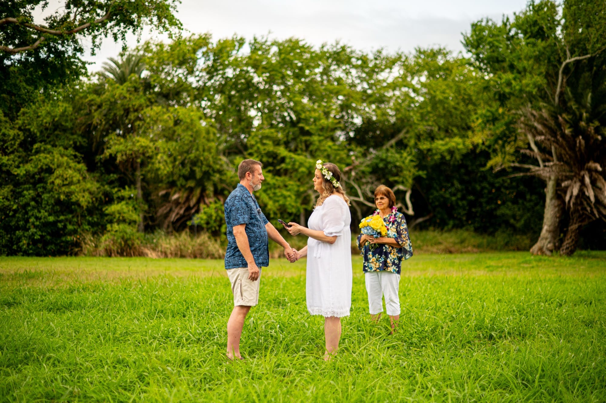 A couple holding hands as they say their vows on the lawn at Kualoa, the officiant beside them