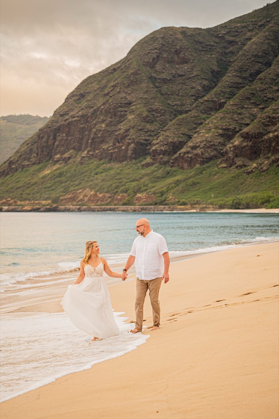 A couple walking the shoreline on the North Shore of Oahu, the mountains behind